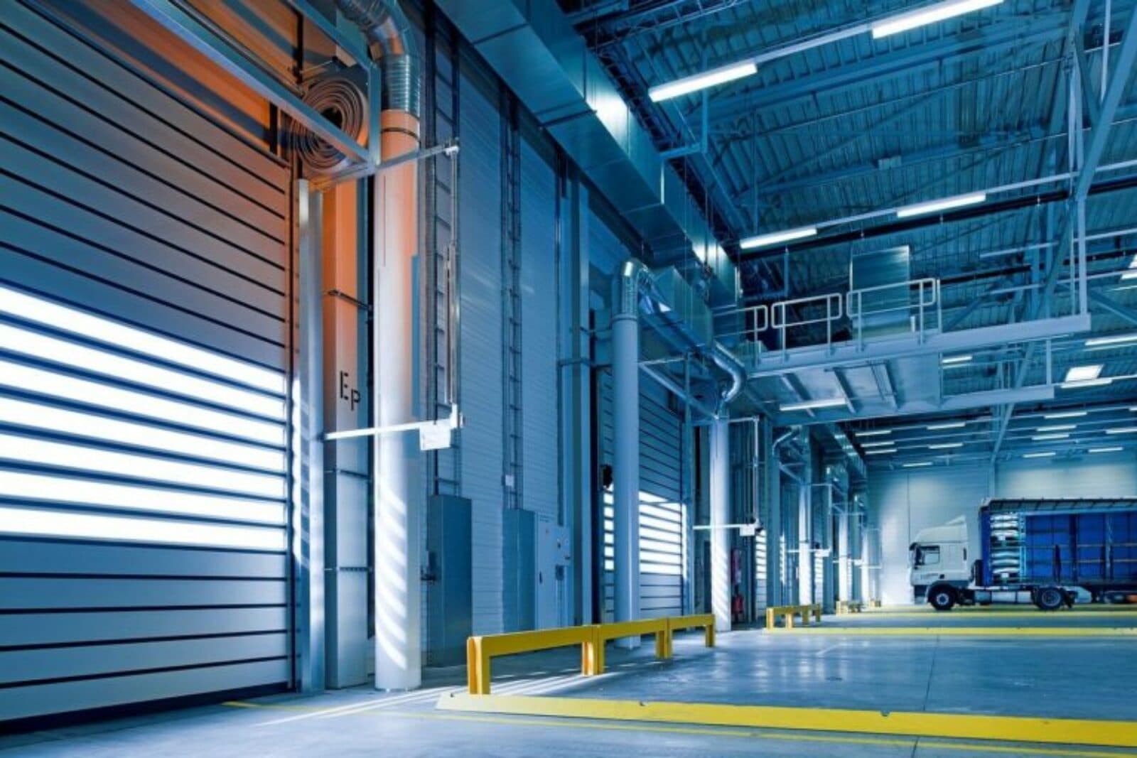 Modern industrial warehouse interior with blue lighting, loading docks, and a parked white semi-truck.