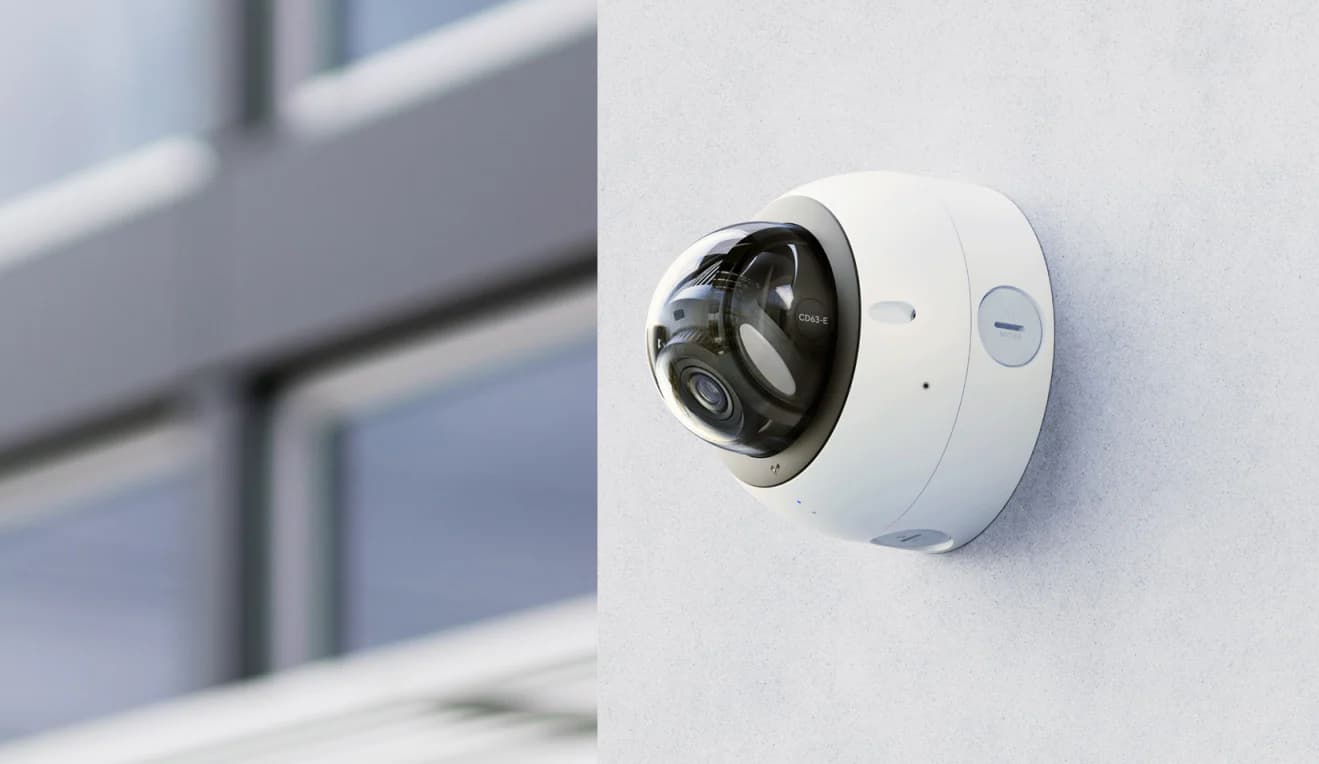 White dome security camera mounted on a light wall with a blurred building background.